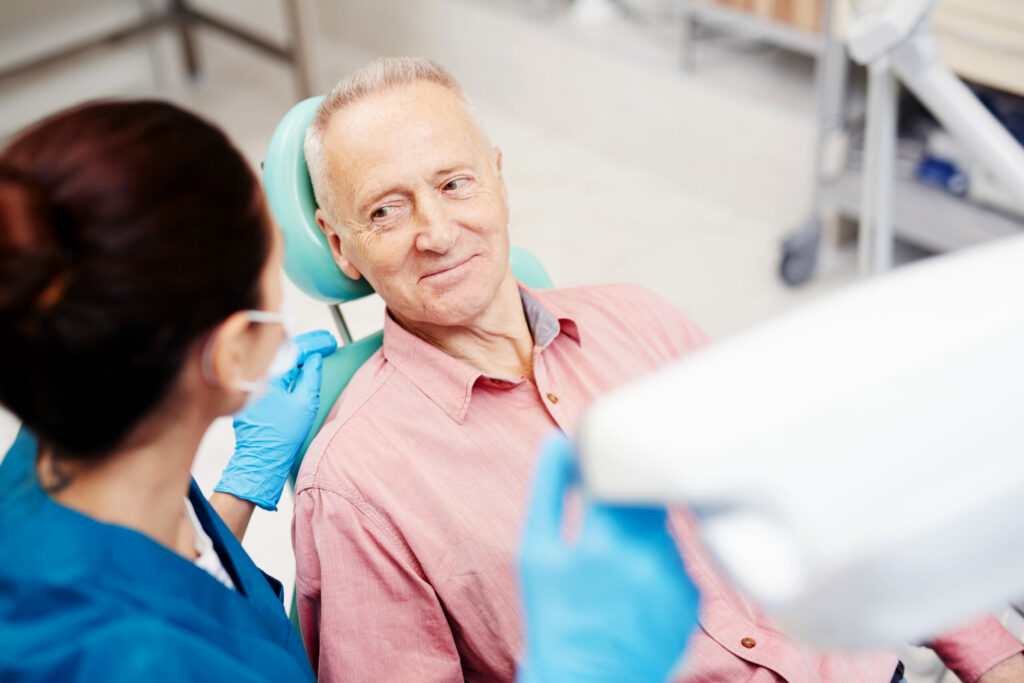 Elderly Man Sitting in Dentist Chair Looking Up at Female Dentist.