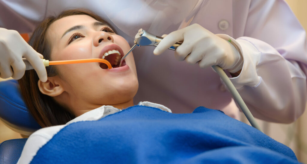 Woman Sitting In Dental Chair With Dentist Behind Her Using Suction and Air Tool
